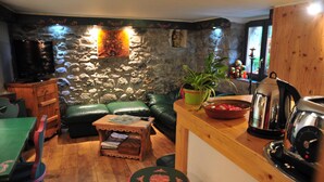 A cozy living room with a stone wall, a green leather sofa, a wooden coffee table, and a kitchen area with a kettle and a coffee pot. - Hôtel du Clocher (Chamonix-Mont-Blanc)