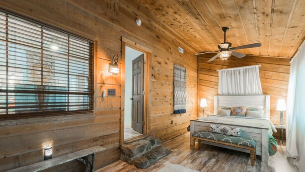 Ein gemütliches Schlafzimmer in einer Blockhaus-Hütte mit einem Bett, einem Deckenventilator und einem Kamin aus Stein.