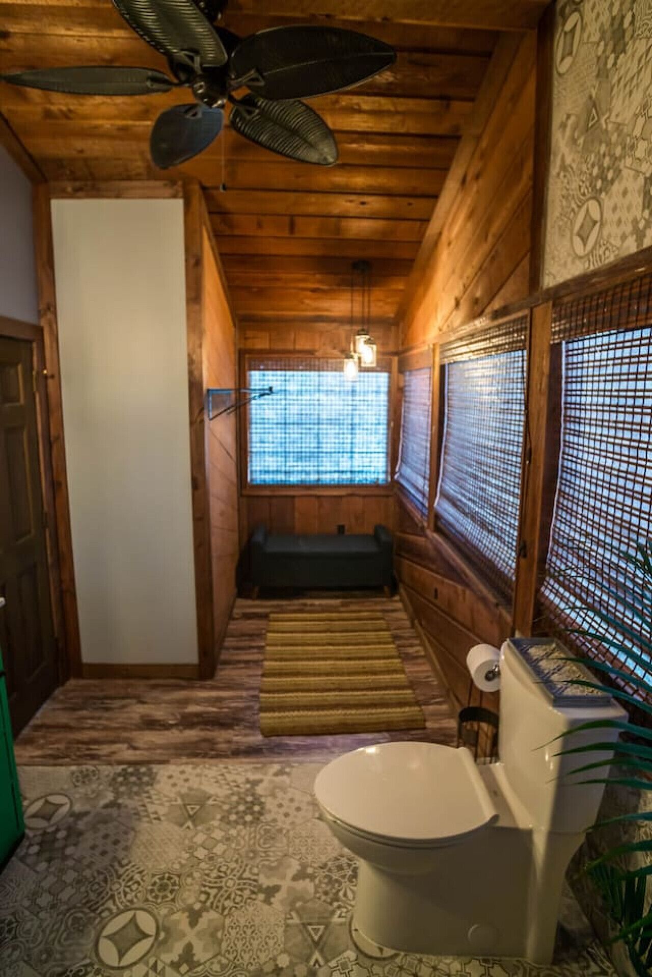 Une salle de bain rustique, aménagée dans une cabane en bois, avec des toilettes, un banc et un ventilateur de plafond.