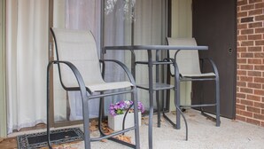 Uma área de pátio com duas cadeiras de metal e uma mesa, uma porta e uma janela com cortinas.