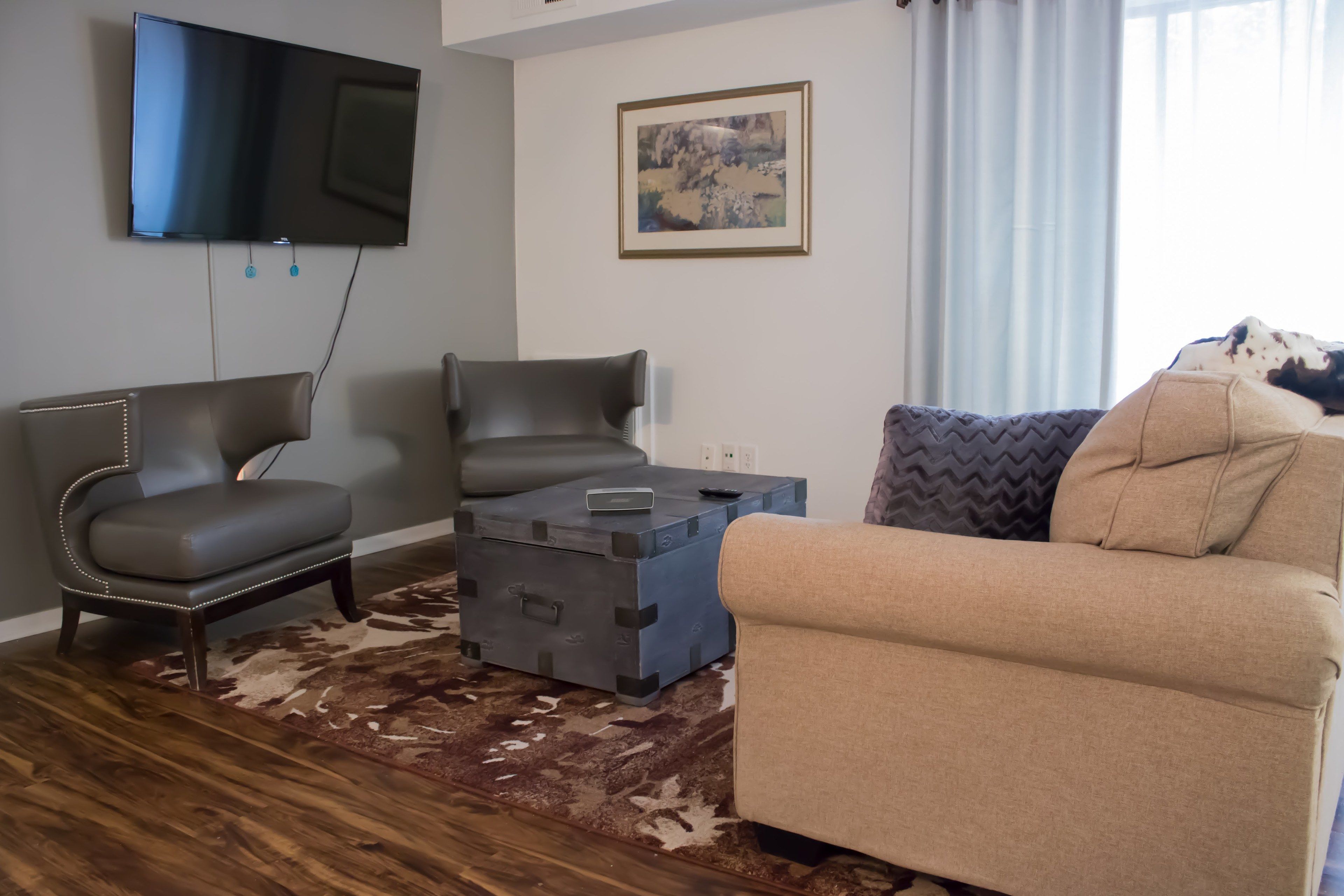 A living room with a flat-screen TV, two leather chairs, a blue storage chest, a beige sofa, and a patterned rug.