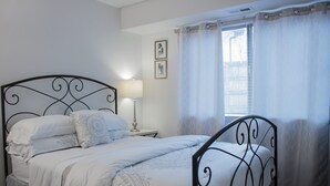 Uma cama bem arrumada com estrutura de metal, um abajur e uma janela com cortinas leves.