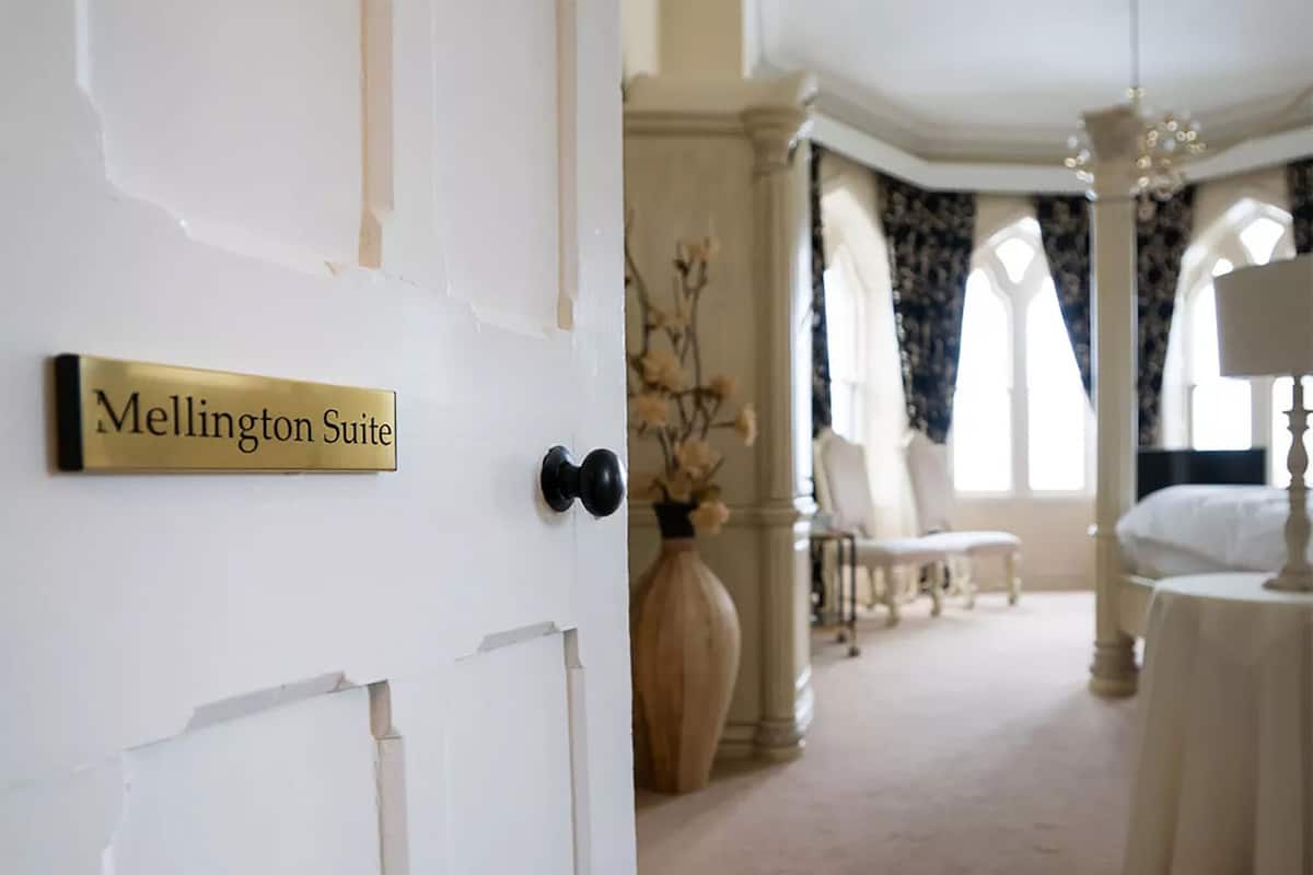 A hotel room with a sign reading "Mellington Suite," a large vase with dried flowers, a bed with white linens, and a chandelier.