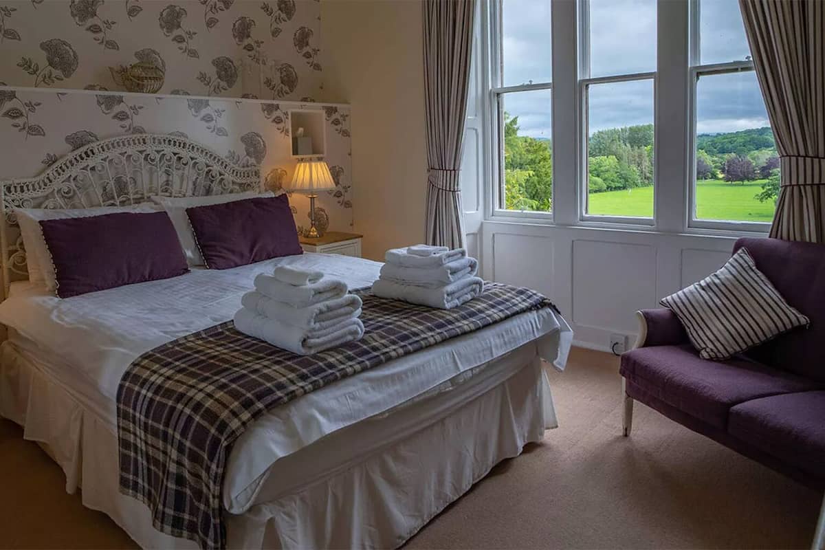 A bedroom with a large bed, two bedside lamps, a headboard with a floral pattern, a window with a view of greenery, and a purple armchair.