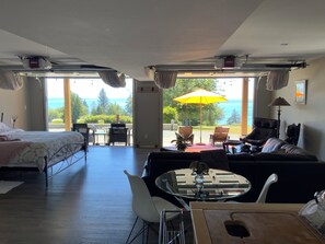 A spacious room with a bed, a sofa, a dining table, and a view of the sea. - Sea of Serenity & Breathtaking Ocean Views & Park-like 6 Acres (Sooke)