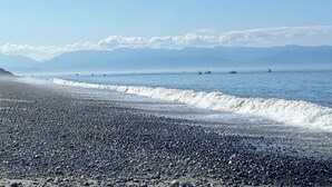 A beach with pebbles, calm water, boats, and mountains in the distance.