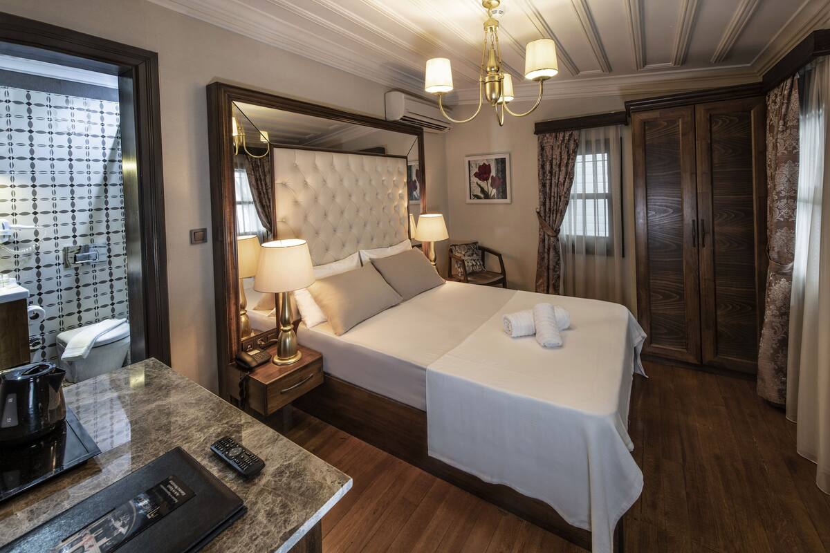 A hotel room with a large bed, a bedside table with a lamp, a small table with a remote control, and a bathroom visible through a glass door.
