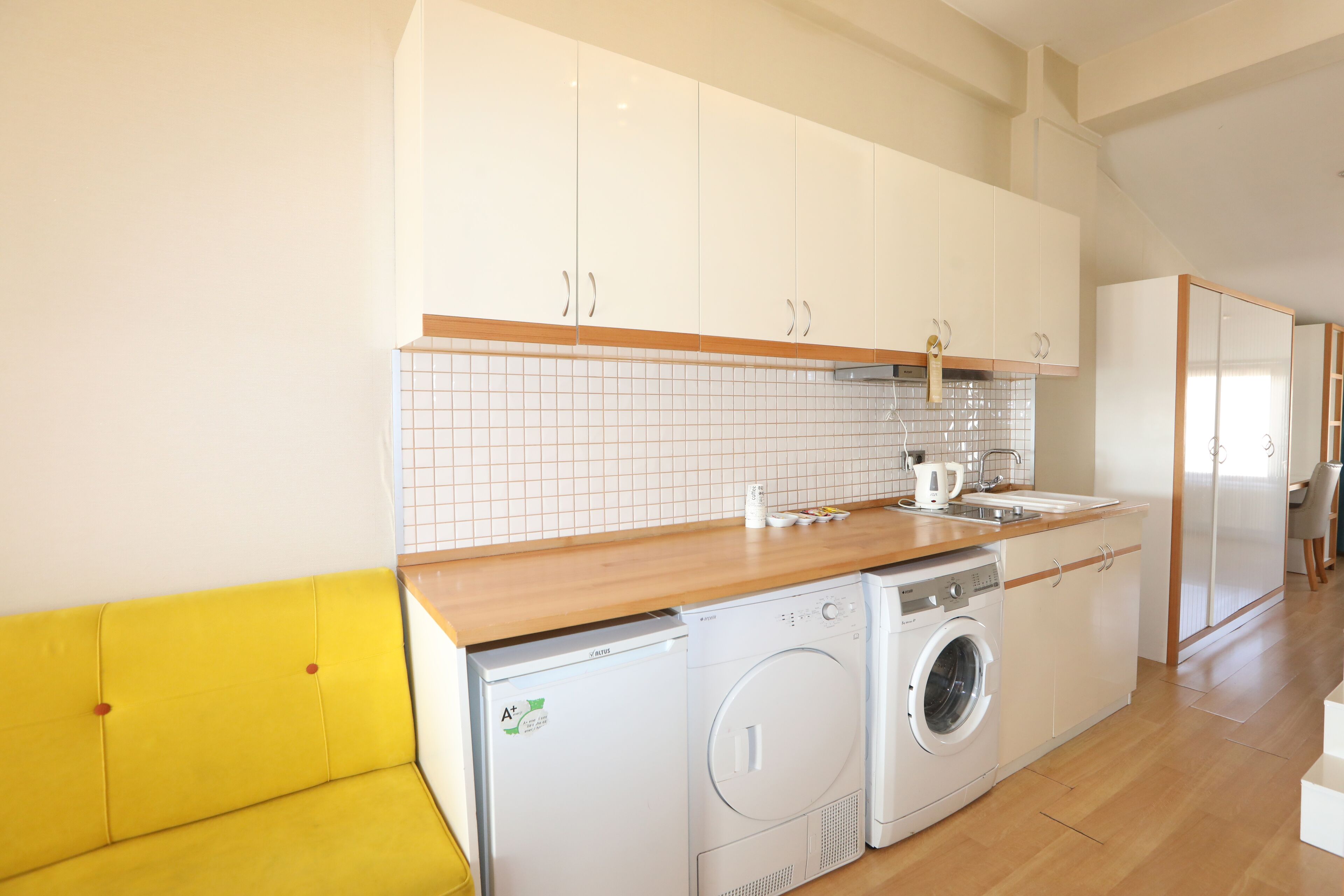 Une cuisine de taille réduite avec des meubles blancs, un plan de travail en bois, un lave-linge et un banc jaune.
