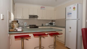 Una cocina con armarios blancos, microondas, estufa y refrigerador. Hay tres taburetes rojos y un jarrón grande en la encimera.