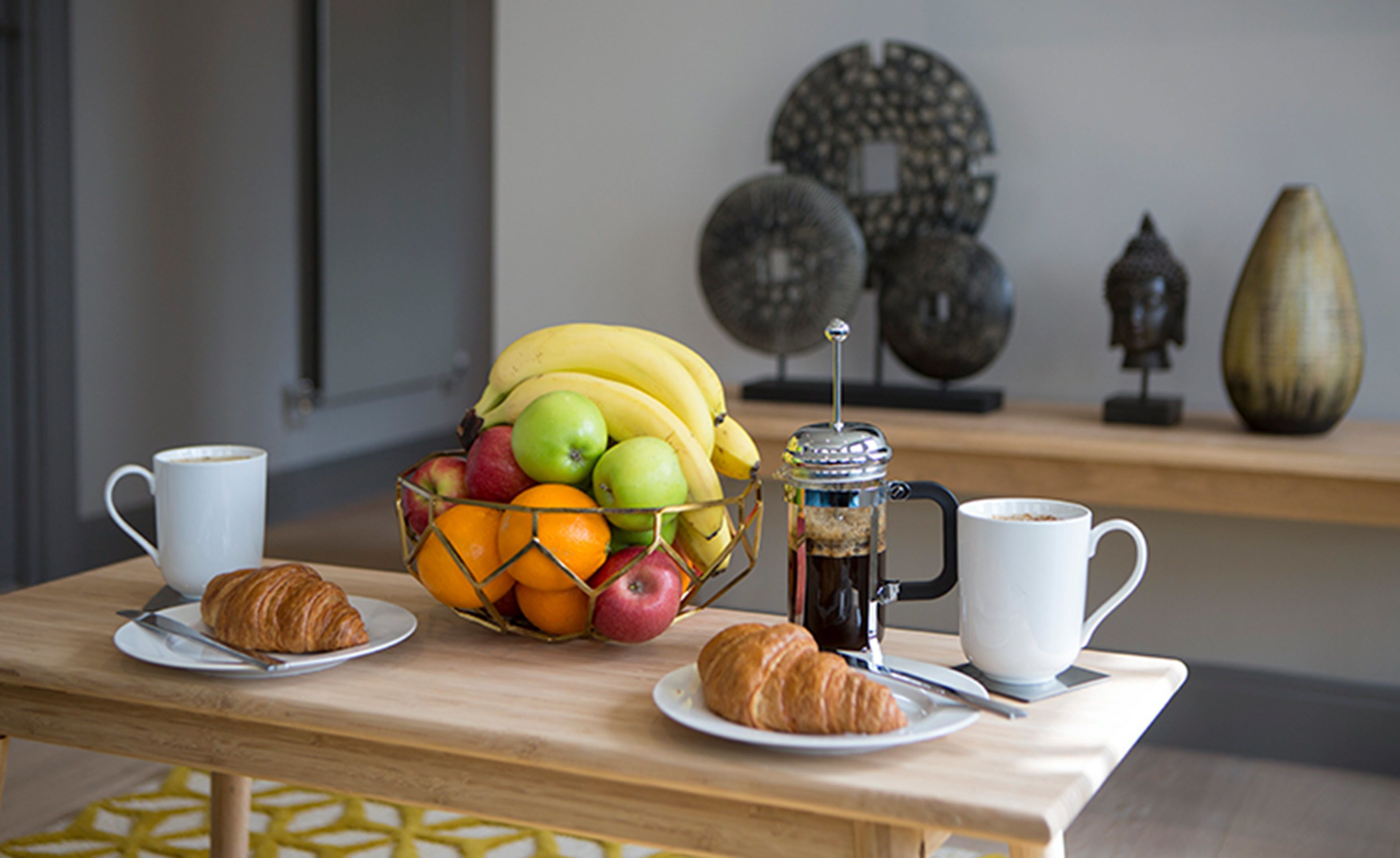 A breakfast setup with croissants, a fruit bowl, and a coffee pot on a wooden table.