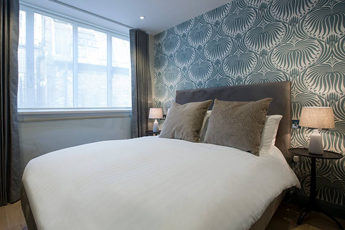 A large bed with a headboard, two bedside tables with lamps, a window with curtains, and a patterned wall.