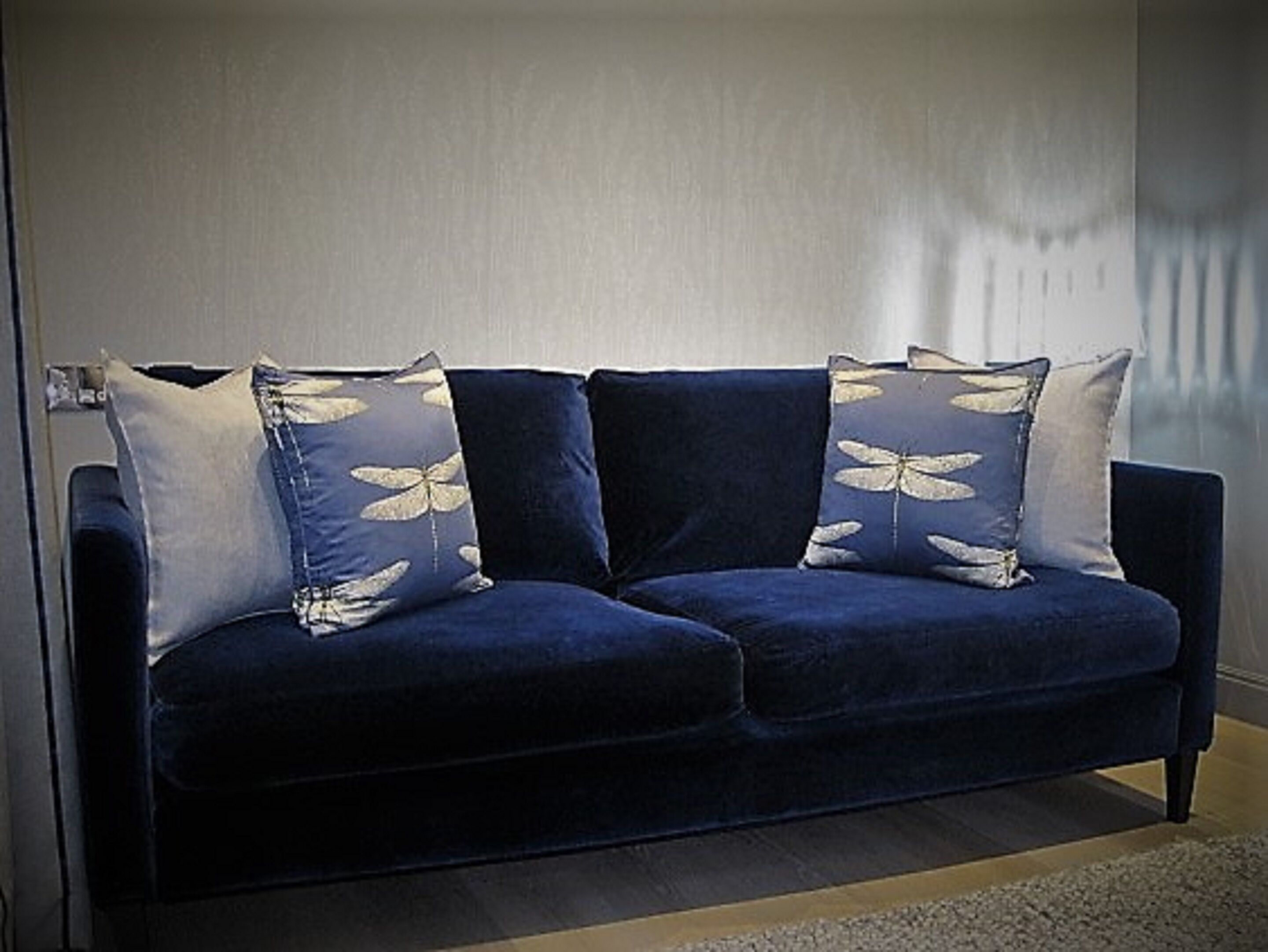 A dark blue sofa with three matching pillows, a light-colored wall, and a wooden floor.