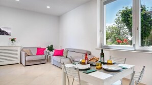 A modern living room with a dining area, featuring a white table set for two, a grey sofa with pink cushions, and a window with a view of trees and flowers.