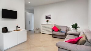 A modern living room with a flat-screen TV, a white console table with a laptop and wine, a beige sofa with red cushions, a white cabinet, and a potted plant.