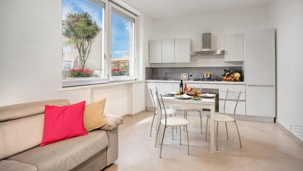 A modern kitchen with white cabinets, a dining table with chairs, and a window with a view of trees and buildings.