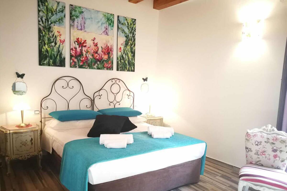A bedroom with a bed, two bedside tables, a chair, and three paintings on the wall.