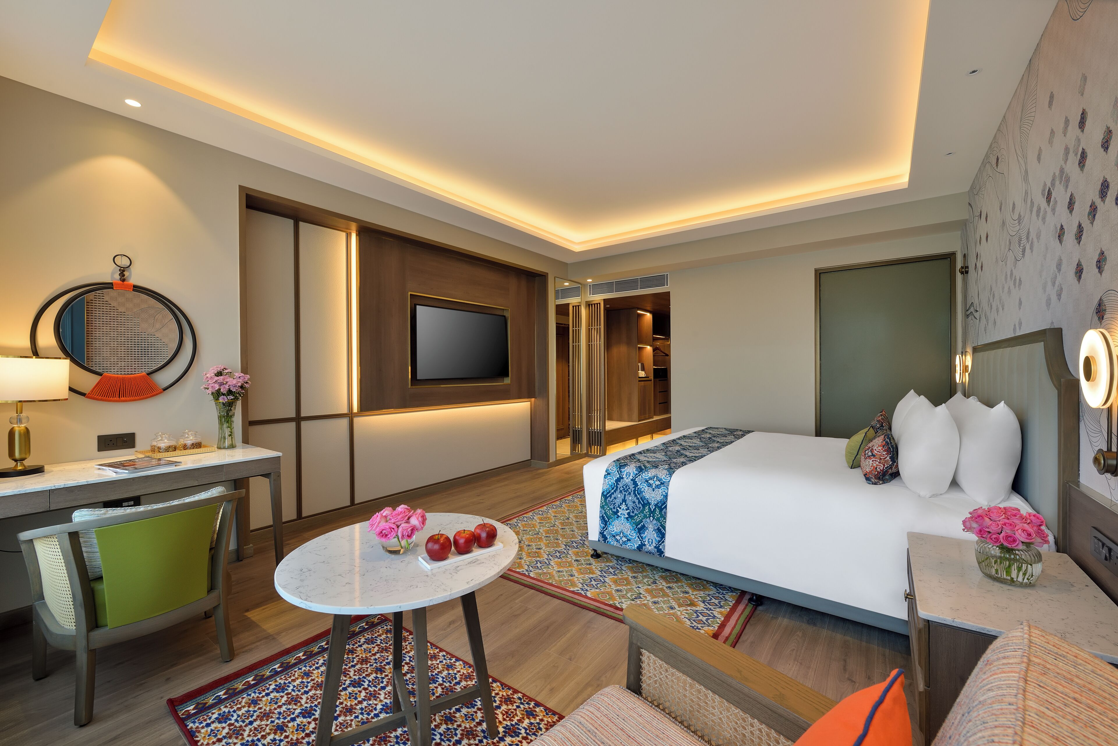 A hotel room with a bed, a desk, a TV, a chair, and a small table with flowers and apples.