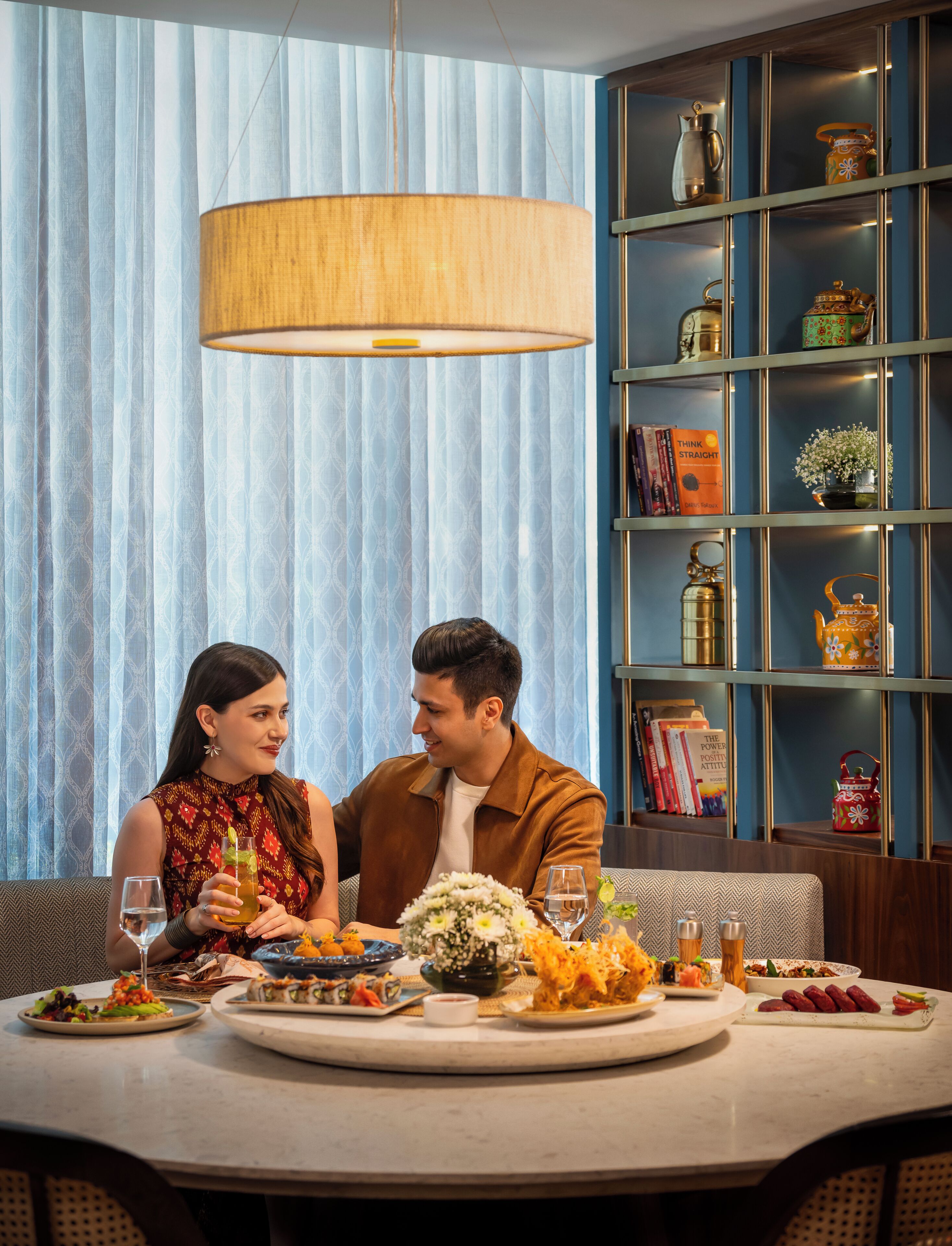 Una pareja cenando en una mesa con diversos platos, una estantería con libros y objetos decorativos, y una lámpara colgante grande.