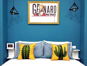 Une chambre avec un mur bleu, une affiche encadrée, deux lampes de chevet et deux coussins ornés de motifs de cactus.