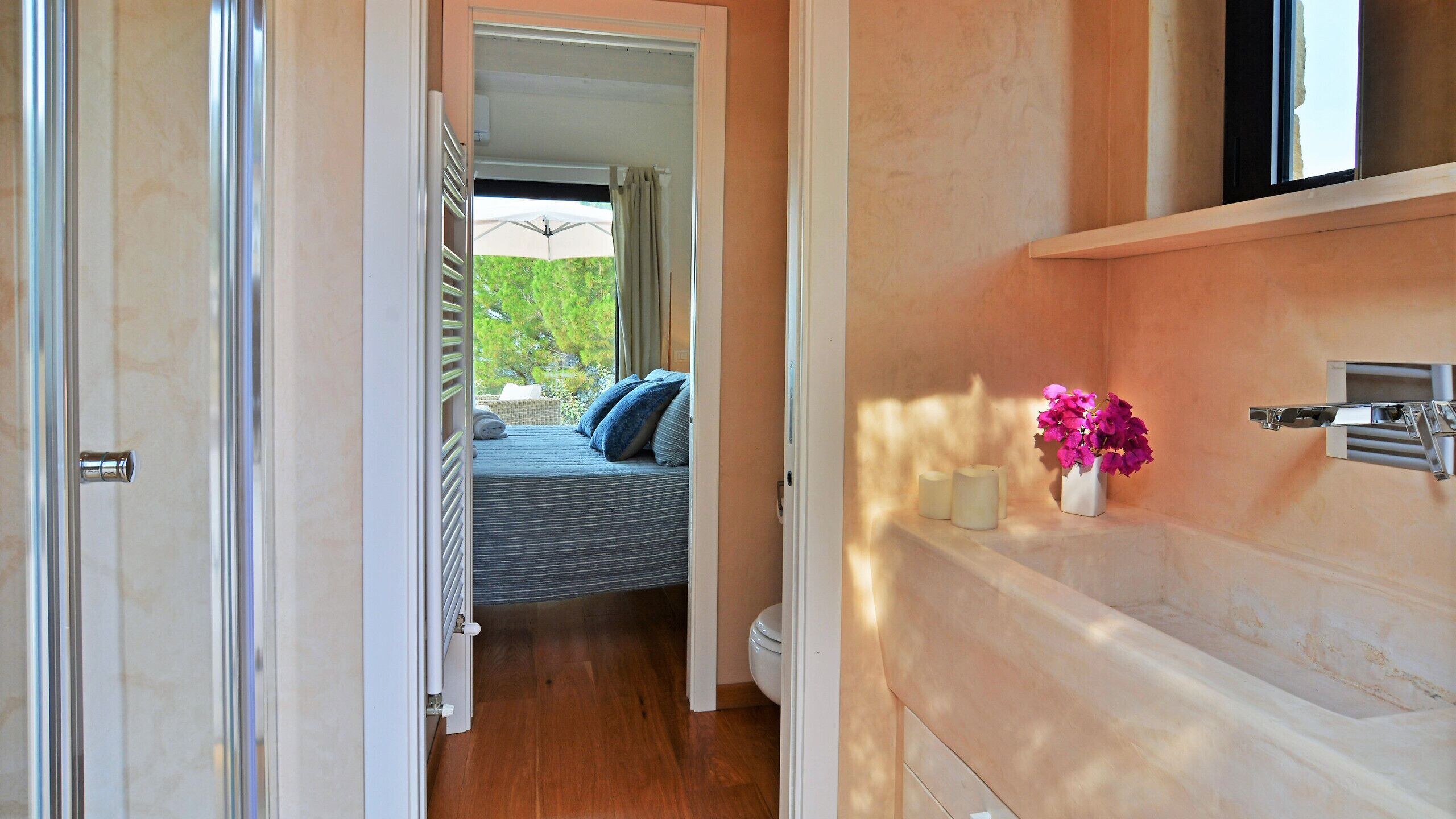 A bedroom with a bed, a window with a view of greenery, a bathroom with a toilet, and a shelf with a vase of flowers.