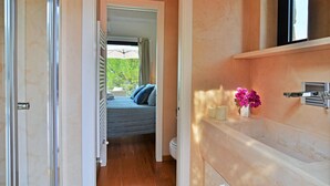 Ein Schlafzimmer mit Bett, einem Fenster mit Blick auf Grünflächen, einem Badezimmer mit Toilette und einem Regal mit Blumen in einer Vase.
