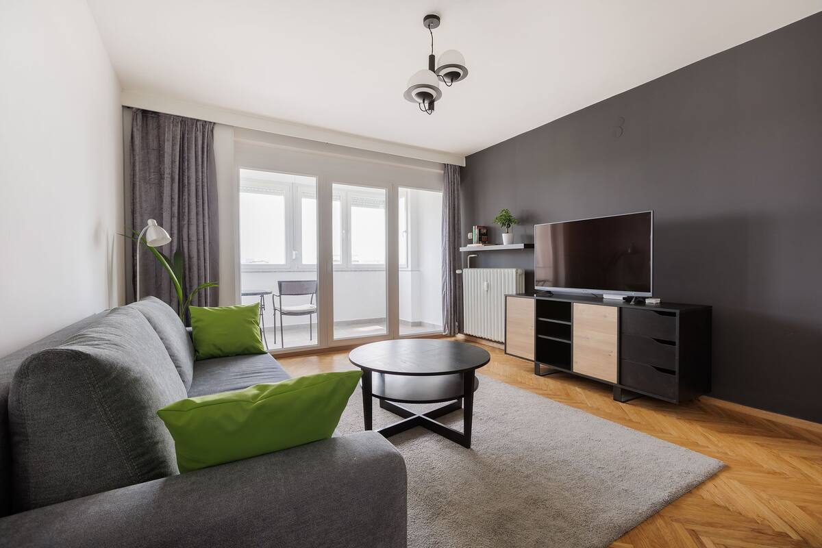 A modern living room with a grey sofa, green cushions, a round coffee table, a TV on a black stand, and a large window with curtains.