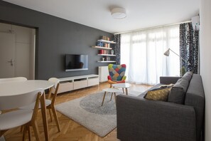 A modern living room with a grey sofa, a colorful armchair, a flat-screen TV, a bookshelf, and a dining area with a white table and wooden chairs.
