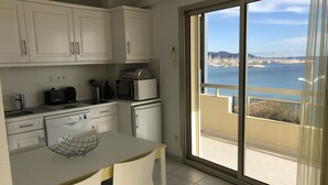 A modern kitchen with white cabinets, a dining area with two chairs, and a large sliding glass door leading to a balcony with a view of the sea and boats. - Superb STUDIO 4 Pers / On Beach / New Condition / Sea view 180 ° except./ Parking / WIFI / AC (Le Lavandou)