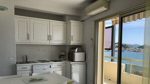 A compact kitchen with white cabinets, a dining table, and chairs. A coffee maker and toaster are on the counter. A sliding glass door leads to a balcony with a view of the sea and boats. - Superb STUDIO 4 Pers / On Beach / New Condition / Sea view 180 ° except./ Parking / WIFI / AC (Le Lavandou)