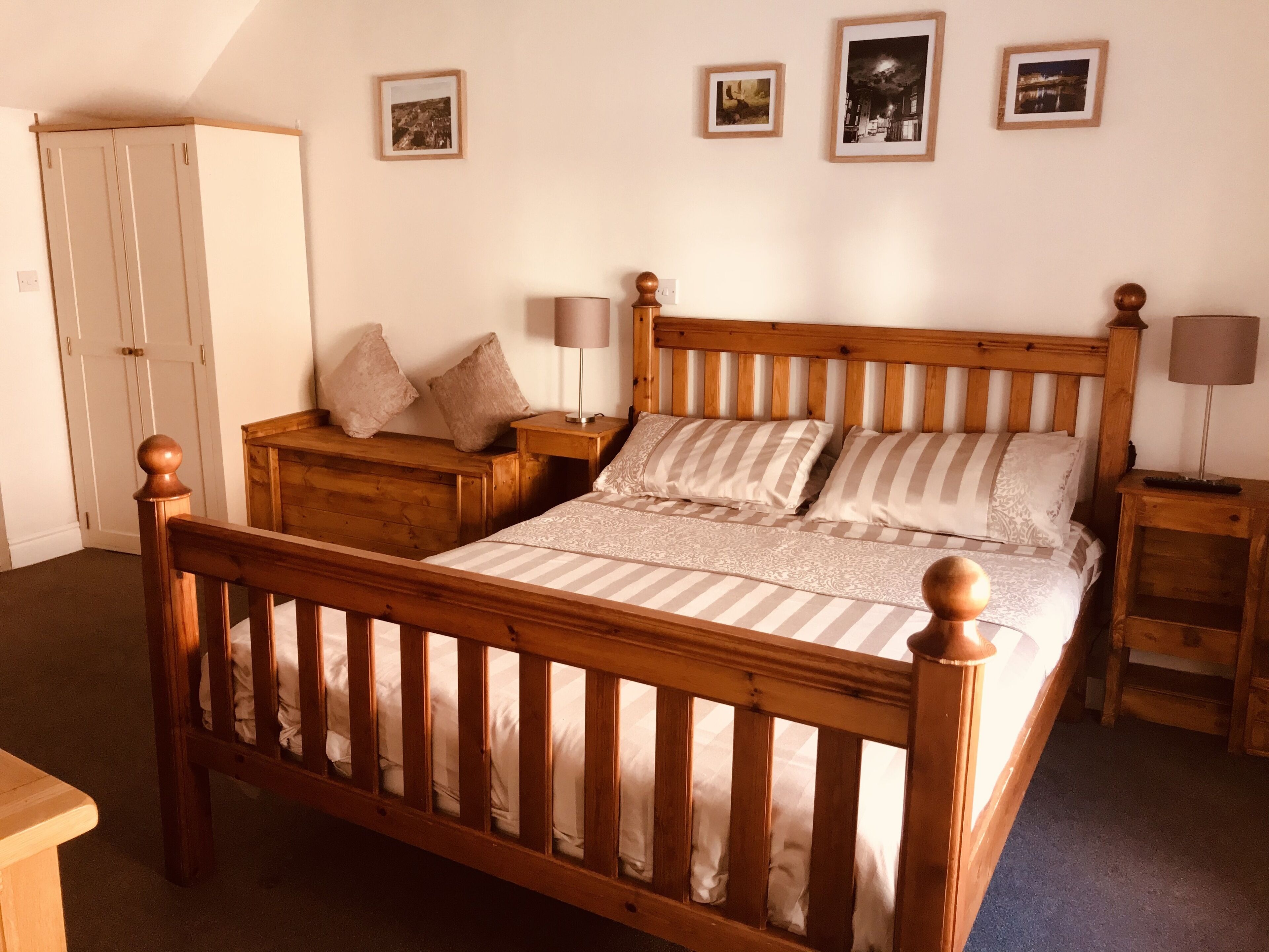 A bedroom with a wooden bed, a nightstand, a bedside table, a wardrobe, and three framed pictures on the wall.
