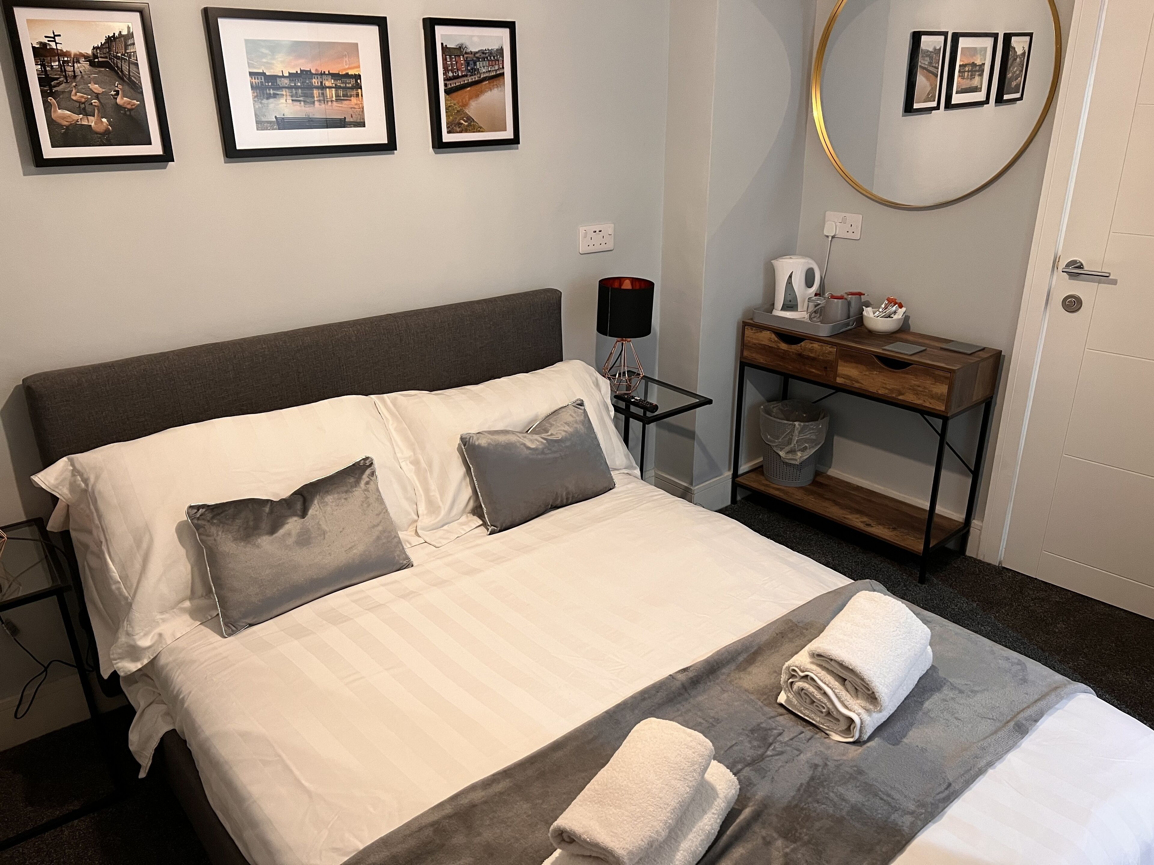 A neatly made bed with pillows, a bedside table with a lamp, a small desk with a mirror, and framed pictures on the wall.