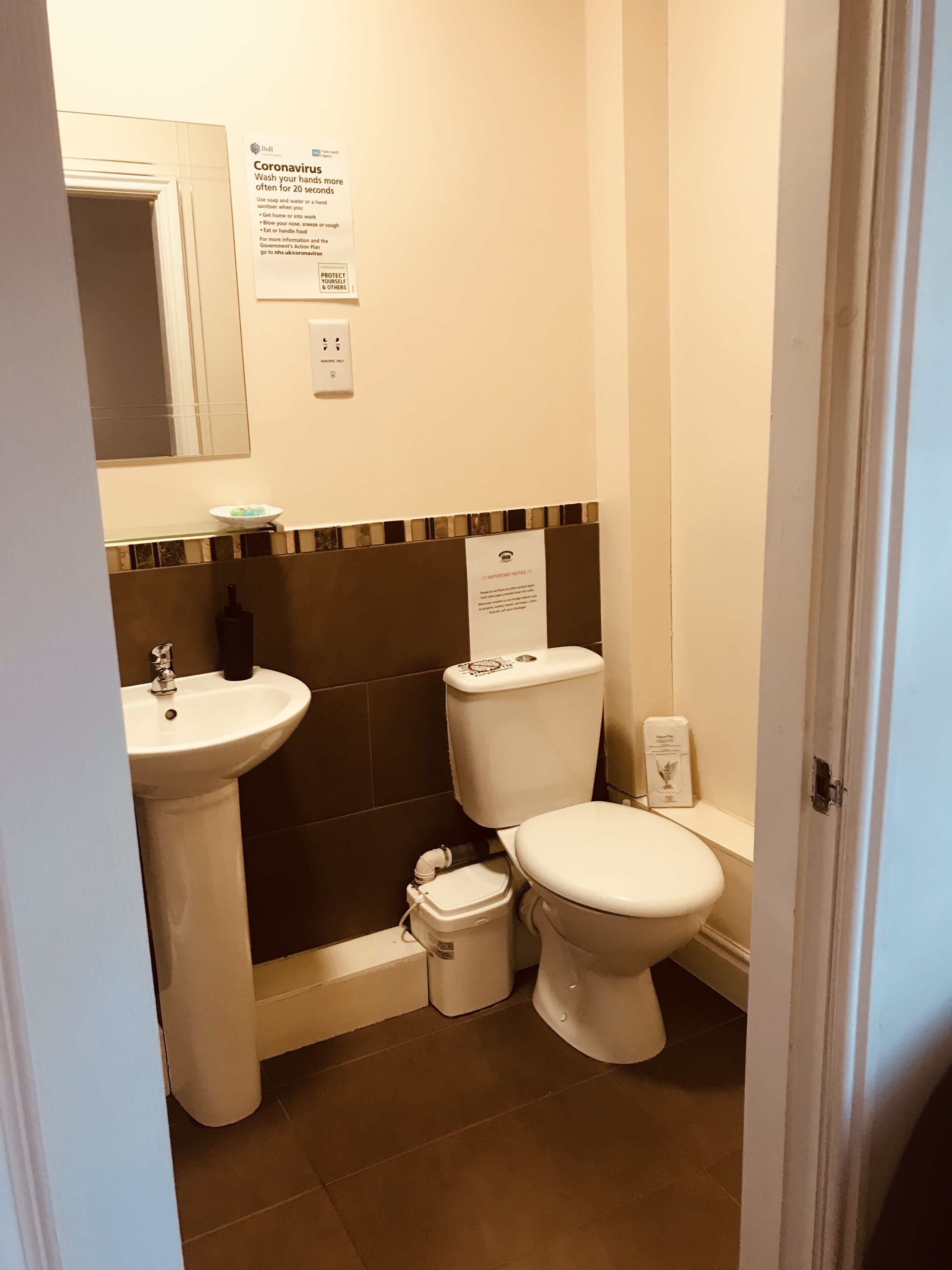 Et lille badeværelse med vask, toilet og bidét. Der er skilte på væggen med information om sikkerhed i forbindelse med coronavirus.