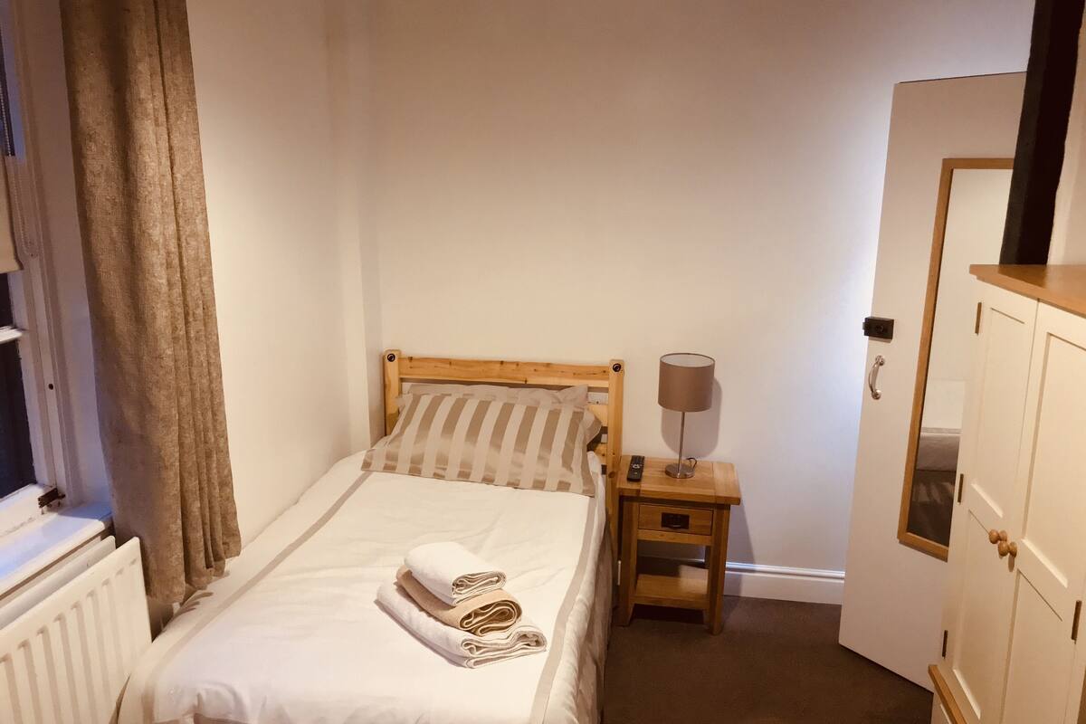 A small, neatly made bed with a wooden headboard, a bedside table with a lamp, and a door to the right.