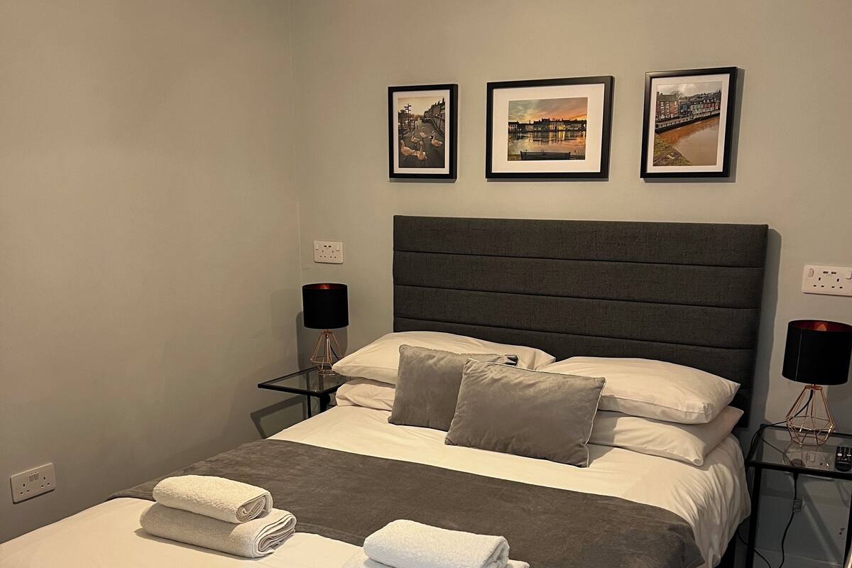 A neatly made bed with white linens and two folded towels, a grey headboard, and three framed pictures on the wall.