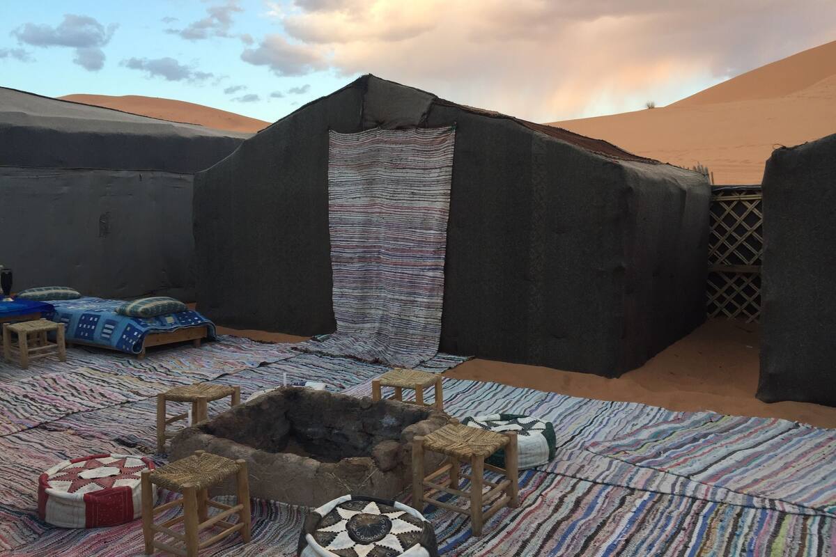 A desert tent with a patterned entrance, a bed with a blue cover, and a fire pit with seating.