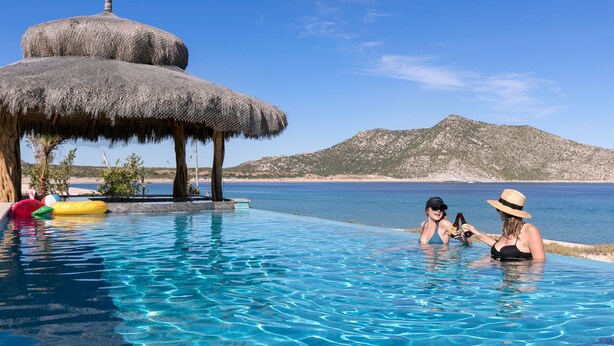 A clear blue pool with a thatched-roof structure, surrounded by a mountainous landscape. - Stunning 6 bedroom home on best sand beach, 45’ pool/jacuzzi, Bar, BBQ, fire-pit (Bahia de Los Frailes)