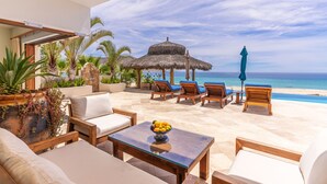 A beachside lounge with a thatched umbrella, wooden lounge chairs, and a view of the ocean. - Stunning 6 bedroom home on best sand beach, 45’ pool/jacuzzi, Bar, BBQ, fire-pit (Bahia de Los Frailes)