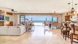 A spacious living area with a large sofa, dining table, and chairs, leading to a balcony with a view of the ocean. - Stunning 6 bedroom home on best sand beach, 45’ pool/jacuzzi, Bar, BBQ, fire-pit (Bahia de Los Frailes)