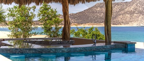 An infinity pool with a thatched roof structure, surrounded by greenery and overlooking a beach and mountains.