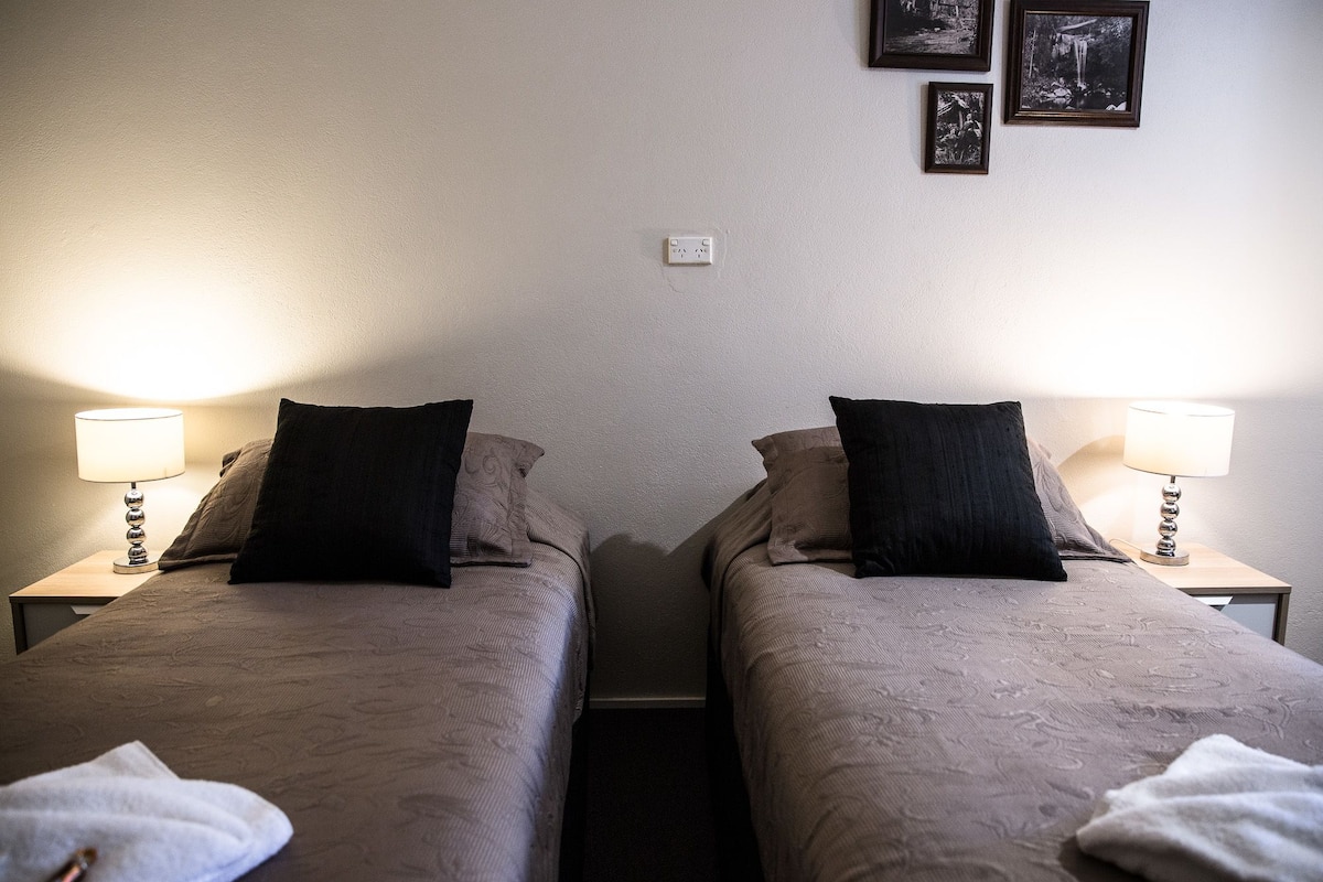 A room with two single beds, each with a bedside table and lamp. There are three framed pictures on the wall.