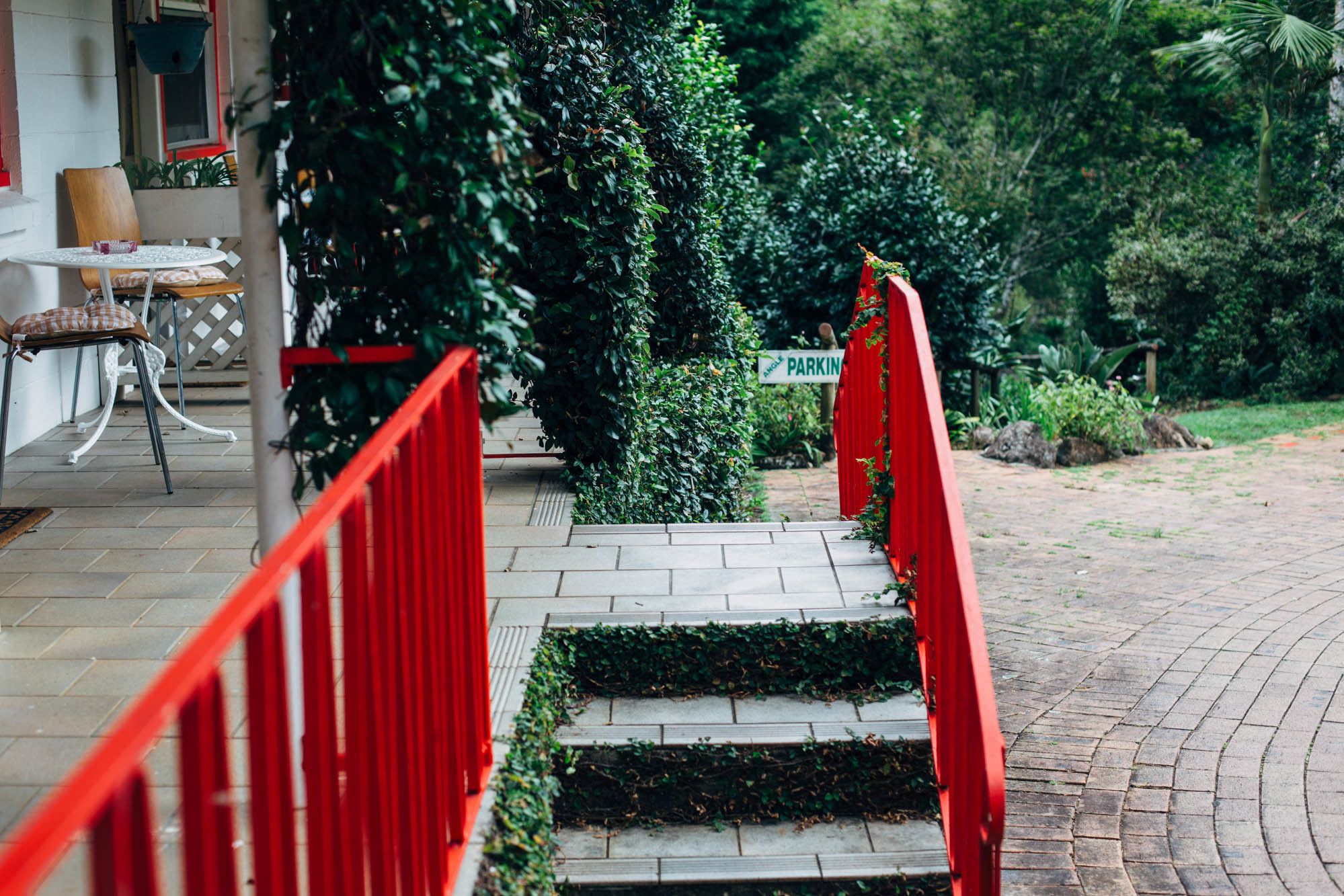 A staircase leading to a parking area with a red railing, surrounded by greenery and a building with a white exterior.
