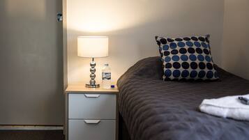 A neatly made bed with a polka dot pillow, a bedside lamp, and a bottle of water on a wooden nightstand with a drawer.
