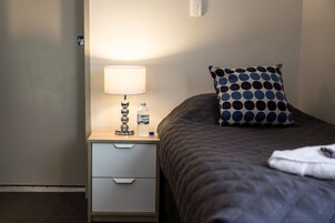 A neatly made bed with a polka dot pillow, a bedside lamp, and a bottle of water on a wooden nightstand with a drawer.