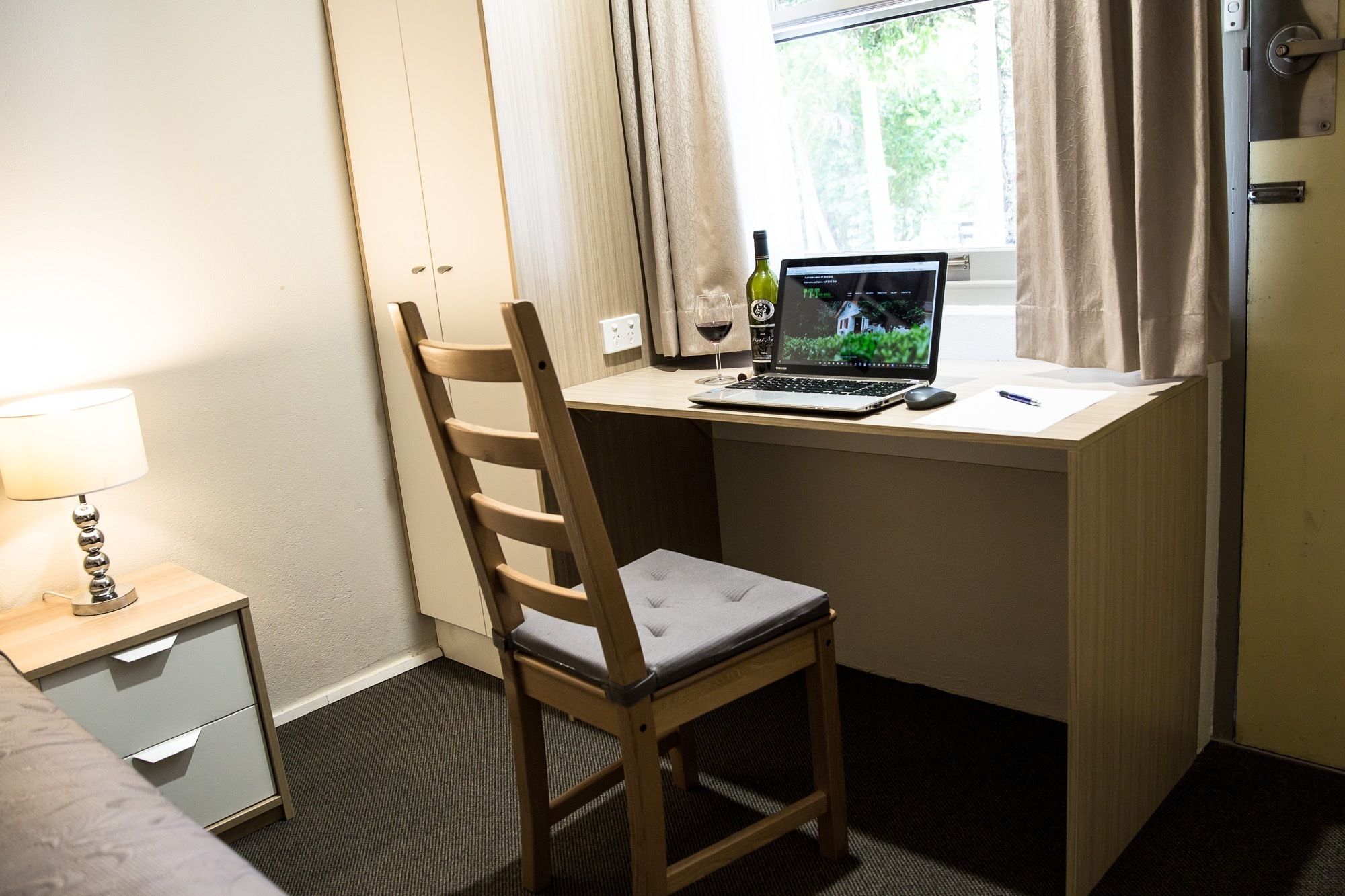 A hotel room with a wooden desk, a laptop, a wine bottle, and a chair.