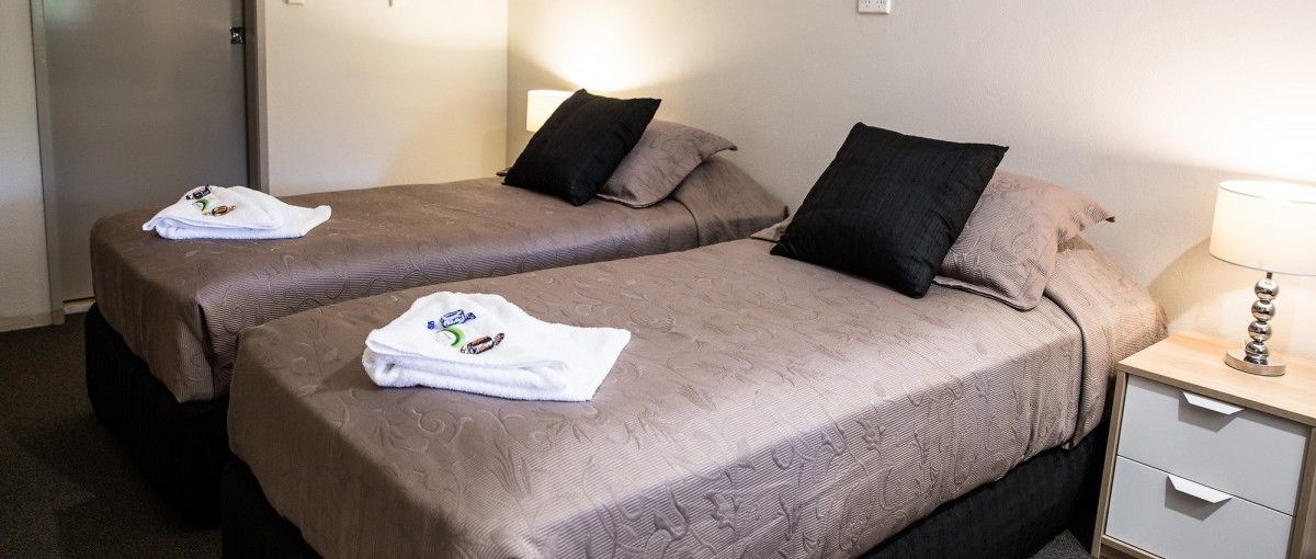 A hotel room with two beds, each with a brown bedspread and a white towel. There is a nightstand with a lamp and a drawer.