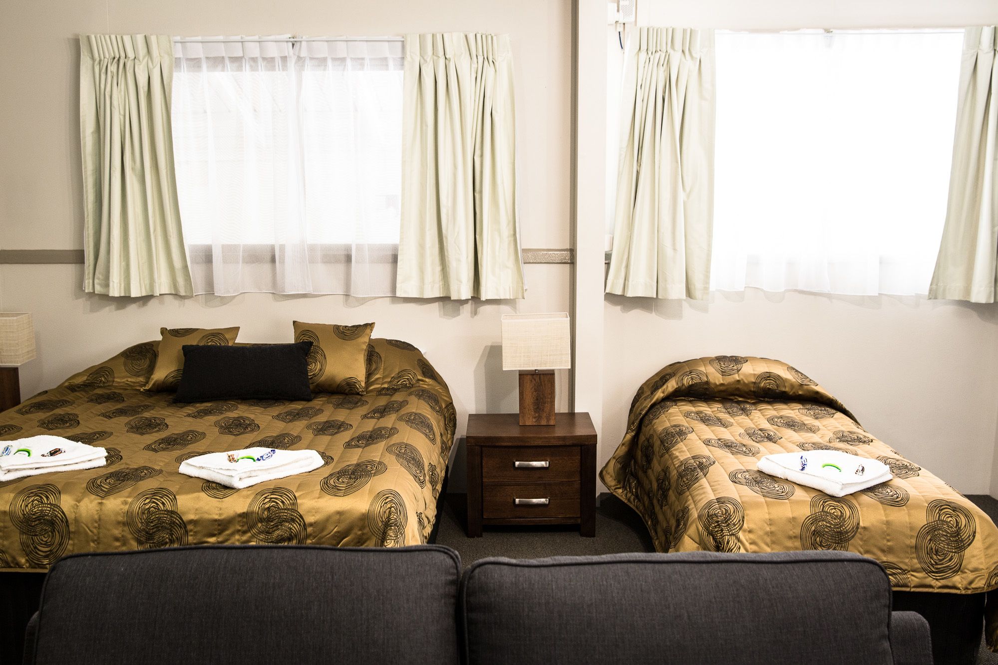 A hotel room with two beds, each with a patterned duvet, a bedside table with a lamp, and curtains on the windows.
