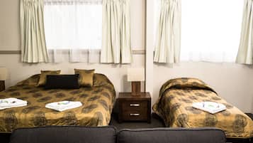 Habitación de hotel con dos camas, cada una con una colcha estampada, una mesita de noche con lámpara y cortinas en las ventanas.