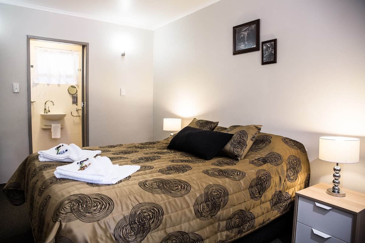 A hotel room with a bed, bedside lamps, a nightstand, a drawer, a bathroom with a sink and mirror, and two framed pictures on the wall.