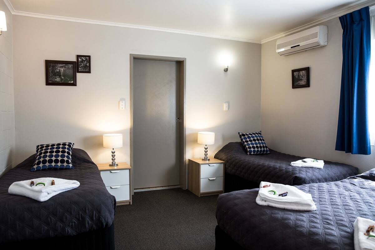 A hotel room with two beds, each with a pillow and a lamp. There is a door in the center, an air conditioning unit on the wall, and a window with curtains.