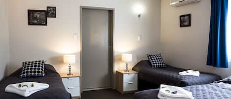 A hotel room with two beds, each with a pillow and a lamp. There is a door in the center, an air conditioning unit on the wall, and a window with curtains.